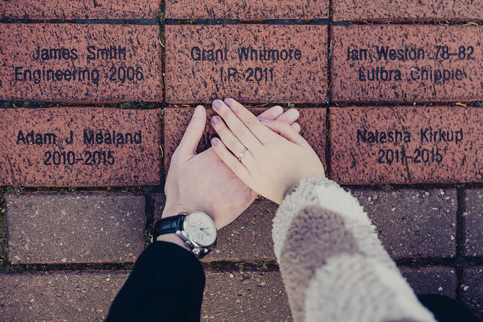 Brown bricks with different names and graduation years written on them with two hands covering a brick together. 